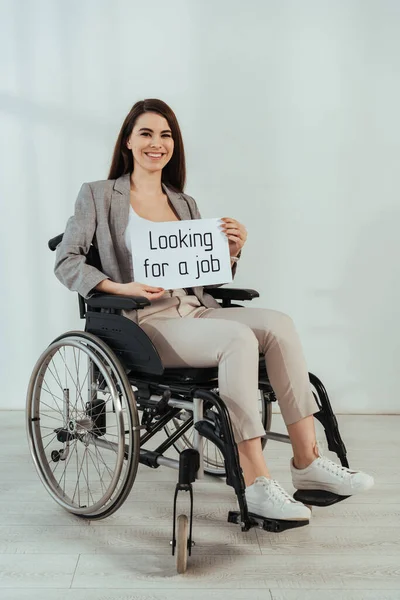 Femme handicapée souriant et tenant une pancarte avec la recherche d'un emploi lettrage sur fauteuil roulant sur blanc — Photo de stock