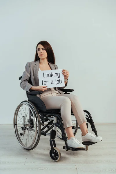 Femme handicapée regardant ailleurs et tenant une pancarte avec la recherche d'un emploi lettrage sur fauteuil roulant isolé sur blanc — Photo de stock