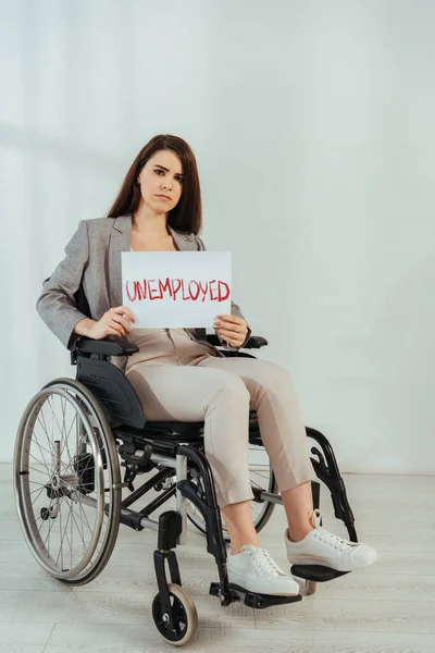 Disabled woman looking at camera and holding placard with unemployed lettering on wheelchair on white — Stock Photo