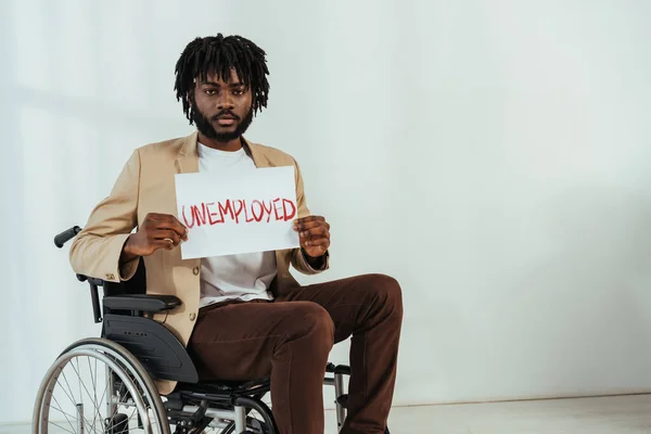 Американский инвалид-американец смотрит в камеру и держит плакат с безработными с надписью на инвалидном кресле на белом — стоковое фото