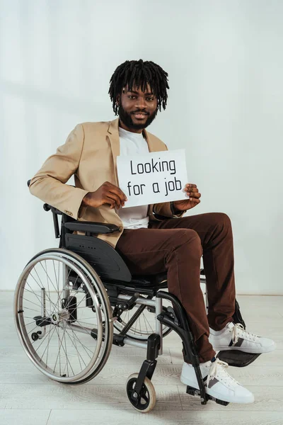Uomo afroamericano disabile con cartello con la ricerca di un lavoro lettering su sedia a rotelle su sfondo bianco — Foto stock