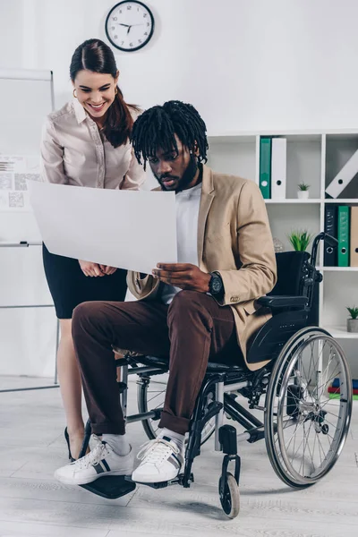 Reclutador sonriendo y mirando papel tejido con afroamericano empleado discapacitado en la oficina - foto de stock