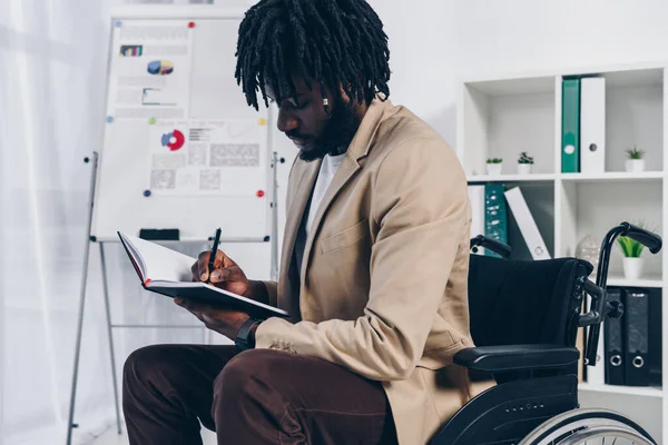 Afrikanisch-amerikanischer behinderter Mitarbeiter im Rollstuhl schreibt im Büro in Notizbuch — Stockfoto