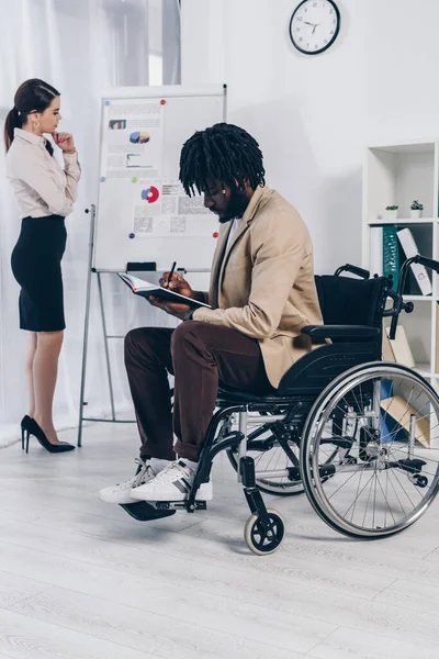 African american disabled employee writing in notebook with thoughtful recruiter near flip chart — Stock Photo