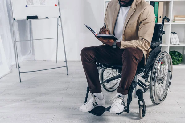 Vista ritagliata del dipendente afro-americano disabile sulla sedia a rotelle scrivendo nel quaderno in ufficio — Foto stock