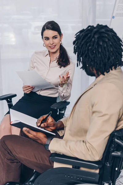 Reclutador con papeles y empleado afroamericano discapacitado con cuaderno hablando en entrevista de trabajo - foto de stock