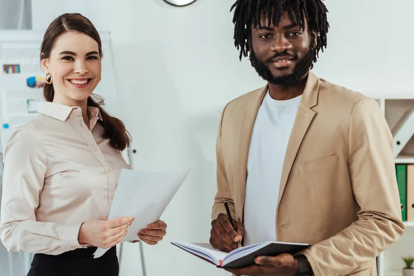 Recruiter with papers and african american employee with notebook looking at camera and smiling — Stock Photo