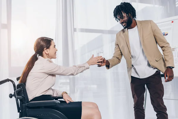 Foco seletivo do funcionário afro-americano dando copo de água para recrutador com deficiência em cadeira de rodas no escritório — Fotografia de Stock