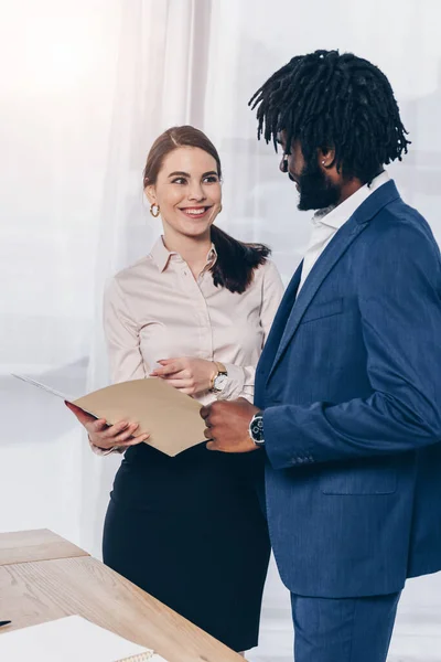 Empleador con la carpeta abierta sonriendo, apuntando a la carpeta abierta y mirando al reclutador afroamericano en la oficina - foto de stock