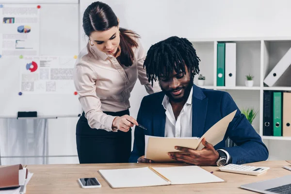 Employé pointant avec stylo vers le dossier ouvert dans les mains masculines afro-américaines au bureau — Photo de stock