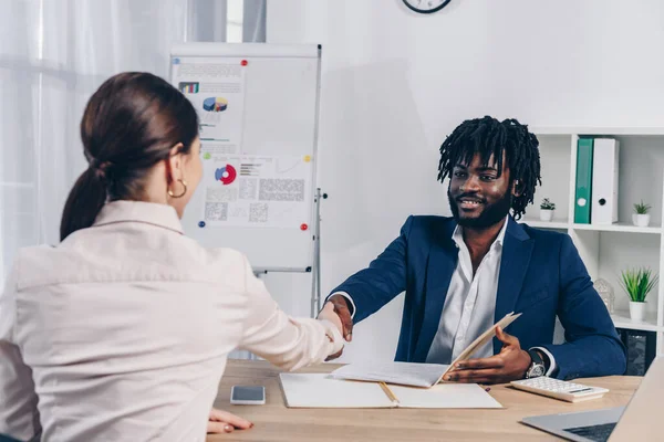 Enfoque selectivo del empleado y reclutador afroamericano mirándose el uno al otro y dándose la mano en la mesa - foto de stock