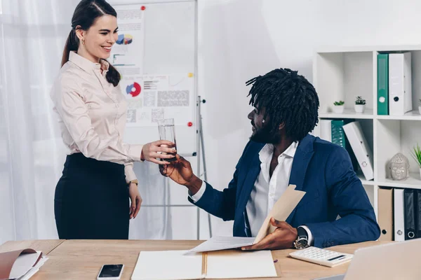 Selective focus of employee getting glass of water to african american recruiter at table in office — Stock Photo