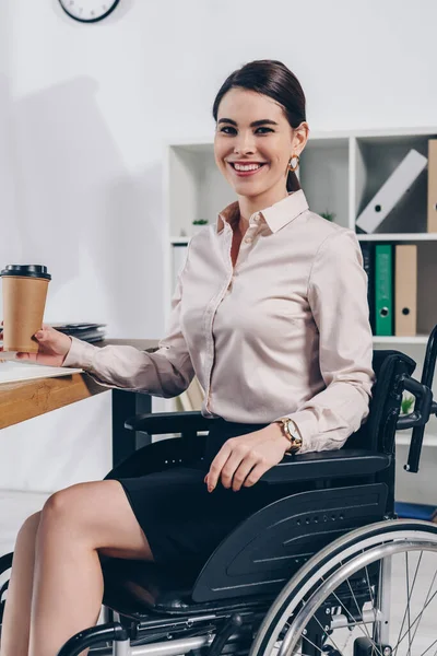 Empleado discapacitado en silla de ruedas mirando a la cámara y sonriendo con una taza de papel de café en la oficina - foto de stock
