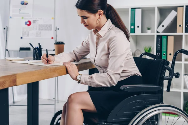 Сотрудник-инвалид на инвалидной коляске пишет в блокноте за столом в офисе — стоковое фото
