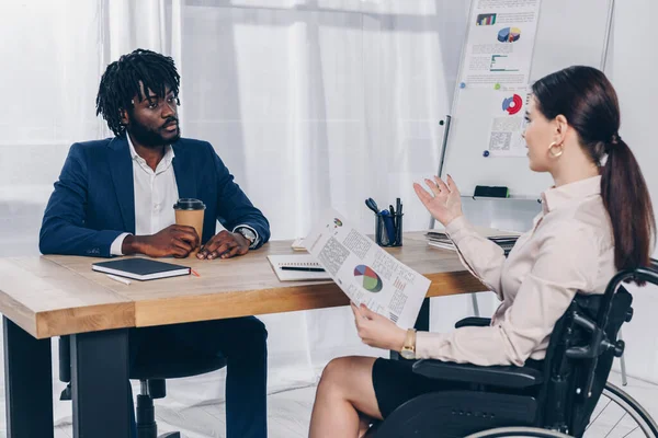 Recrutador afro-americano e empregado com deficiência em cadeira de rodas conversando à mesa no escritório — Fotografia de Stock