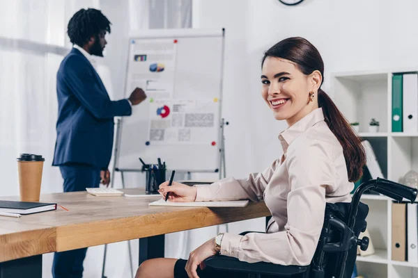 Recrutador afro-americano perto de flip chart e empregado deficiente escrevendo em notebook, sorrindo e olhando para a câmera no escritório — Fotografia de Stock