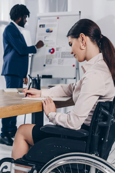 Afrikanischer Rekrutierer in der Nähe von Flipchart und behinderter Mitarbeiter schreibt in Notizbuch — Stockfoto