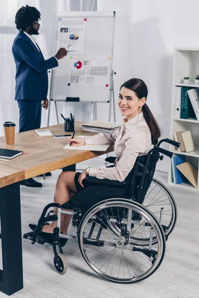 Selektiver Fokus eines afrikanisch-amerikanischen Personalvermittlers in der Nähe von Flipchart und behindertem Mitarbeiter, der in Notizbuch schreibt und in die Kamera blickt — Stockfoto