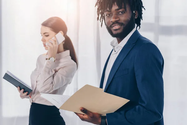 Selective focus of african american employer with folder looking at camera and recruiter talking on smartphone — Stock Photo