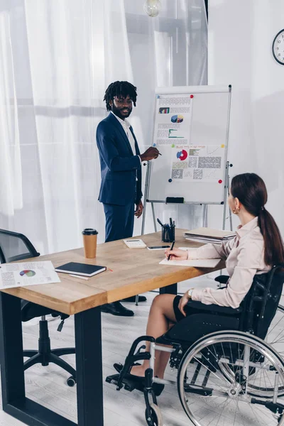 African american recruiter near flip chart looking at disabled employee writing in notebook in office — Stock Photo