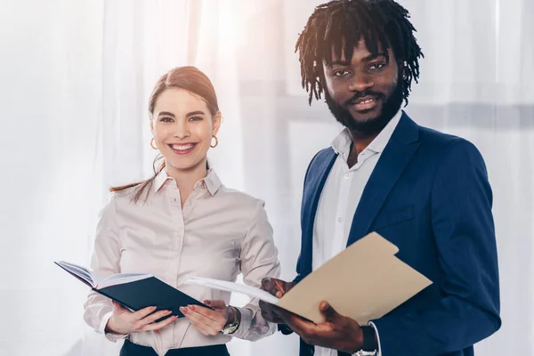 Employeur afro-américain avec dossier et recruteur avec ordinateur portable souriant et regardant la caméra — Photo de stock