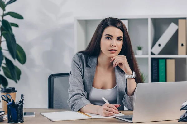 Recruiter looking at camera and writing in notebook near laptop in office — Stock Photo