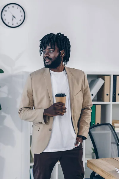 Recruteur afro-américain regardant loin avec la main dans la poche et tasse de papier de café dans le bureau — Photo de stock