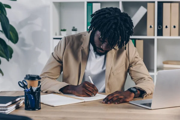 Reclutador afroamericano enfocado en escribir en cuaderno en la mesa en la oficina - foto de stock