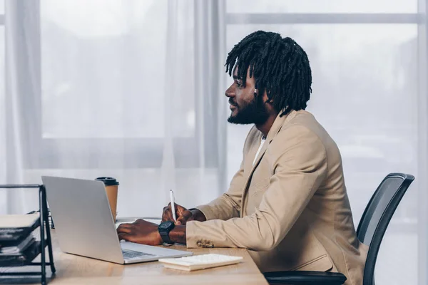 Foco seletivo do recrutador americano africano escrevendo com caneta perto do laptop à mesa no escritório — Fotografia de Stock