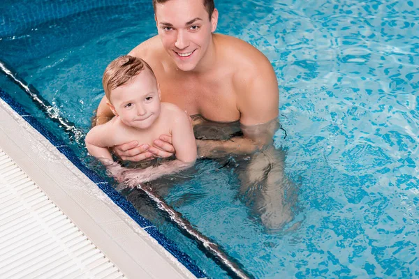 Entraîneur de natation heureux tenant dans les bras tout-petit garçon tout en nageant dans la piscine — Photo de stock