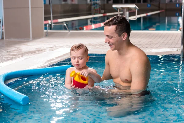 Beau entraîneur de natation souriant tout en regardant tout-petit garçon jouer dans la piscine — Photo de stock
