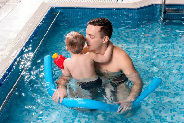 Guapo entrenador de natación nadando con un niño pequeño y sosteniendo fideos de piscina - foto de stock