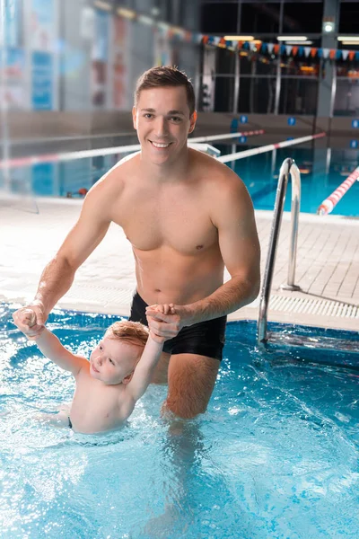 Entraîneur de natation positif tenant la main avec enfant en bas âge dans la piscine — Photo de stock