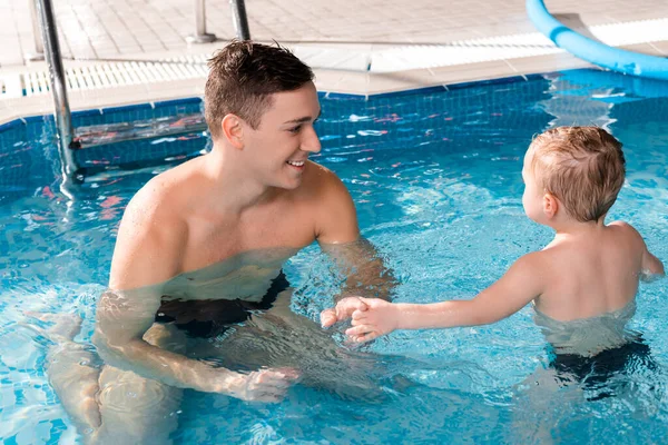 Entrenador de natación positivo cogido de la mano con niño pequeño en la piscina - foto de stock