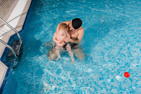 Вид зверху усміхненого тренера з плавання в тренувальній шапці малюка хлопчика в басейні — стокове фото