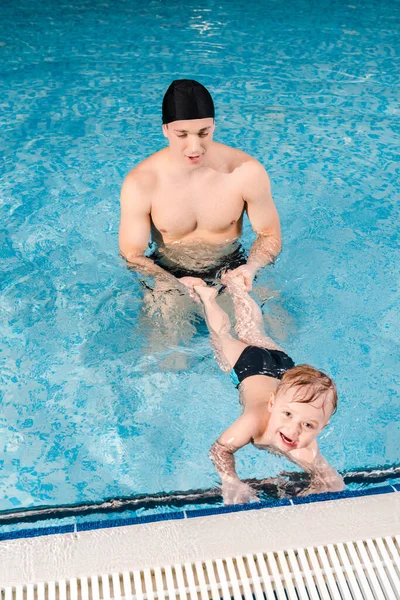 Entrenador de natación en gorra de natación sosteniendo las piernas de niño alegre en la piscina - foto de stock