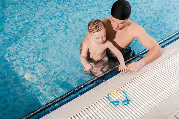 Overhead view of swim coach in swimming cap and toddler boy looking at goggles — Stock Photo