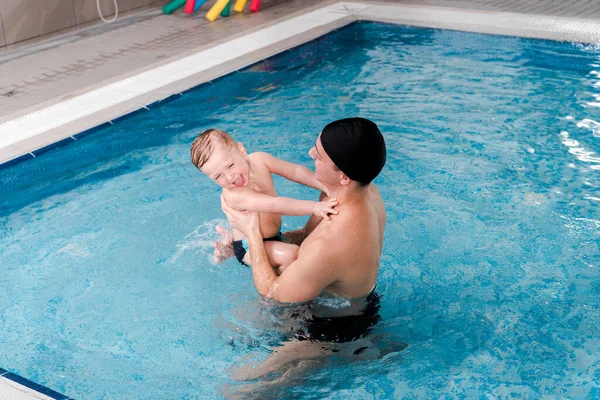 Entrenador de natación feliz en gorra de natación sosteniendo en brazos niño alegre - foto de stock