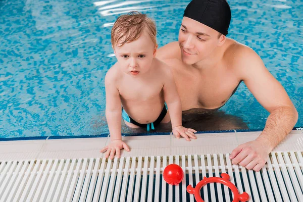 Schöner Schwimmtrainer mit Badekappe schaut Kleinkind im Schwimmbad an — Stockfoto