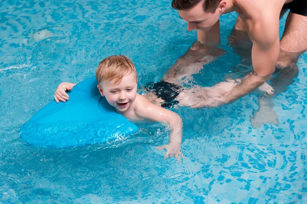 Tout-petit excité garçon nageant avec planche flottante près de l'entraîneur de natation — Photo de stock