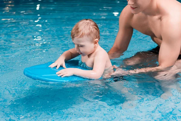 Tout-petit garçon nageant avec planche flottante près entraîneur de natation musculaire — Photo de stock