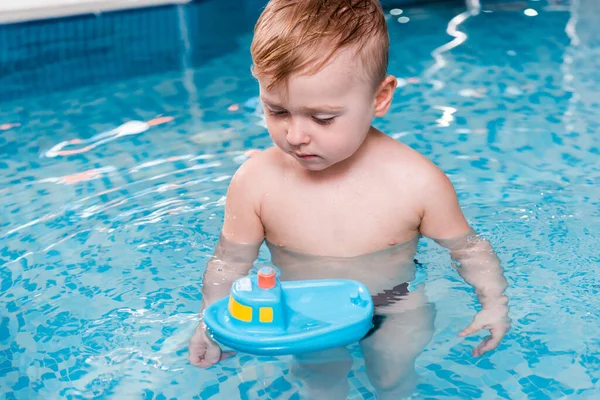 Cute toddler boy looking at toy ship in swimming pool — Stock Photo