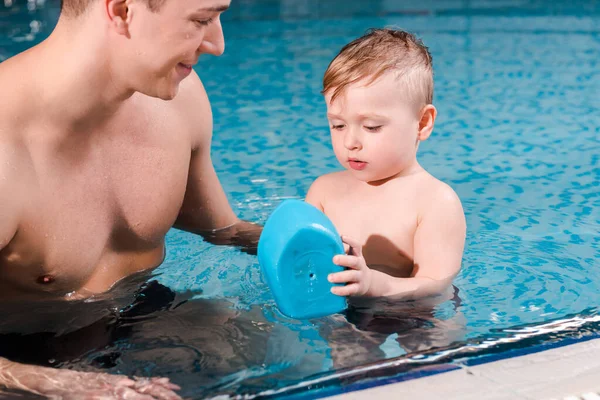 Happy swim coach looking at toddler boy playing with toy ship in swimming pool — Stock Photo