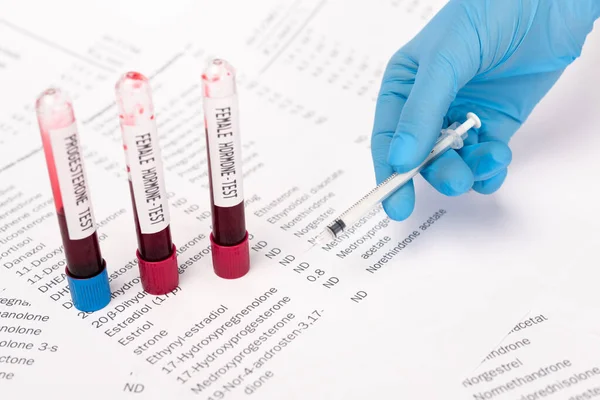 Cropped view of doctor holding syringe near test tubes with blood samples and lists with hormones — Stock Photo