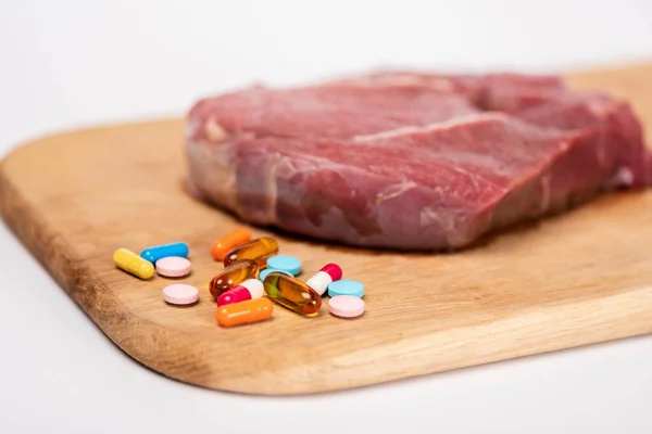 Enfoque selectivo de píldoras hormonales y carne cruda en tabla de cortar de madera aislada en blanco - foto de stock