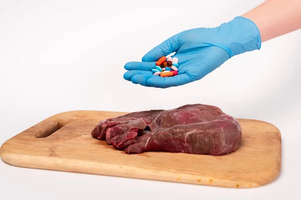 Vista recortada del médico sosteniendo píldoras hormonales cerca de la carne cruda en la tabla de cortar sobre fondo blanco - foto de stock