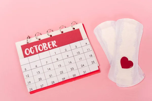 Vista de alto ângulo de almofadas femininas com coração de papel e calendário em fundo rosa — Fotografia de Stock