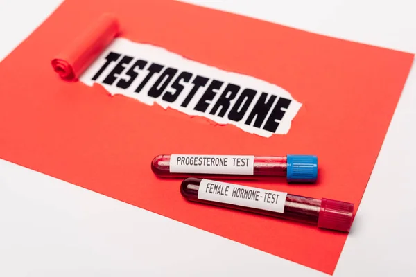 Крупный план пробирки с прогестероном и женскими образцами гормональной крови возле красной бумаги с надписью тестостерона на белом фоне — стоковое фото