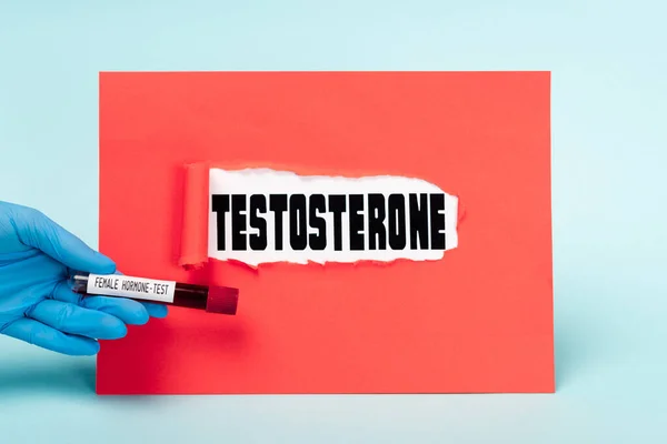 Cropped view of doctor holding test tube with blood sample of female hormone near testosterone lettering in hole of red paper on blue background — Stock Photo