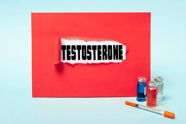 Gläser mit Hormonpräparaten und Spritze in der Nähe von Testosteron-Schriftzug im roten Papierloch auf blauem Hintergrund — Stockfoto